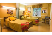Two-bed senior living resident room with yellow bedspreads, bedside lamps, a window and seating.