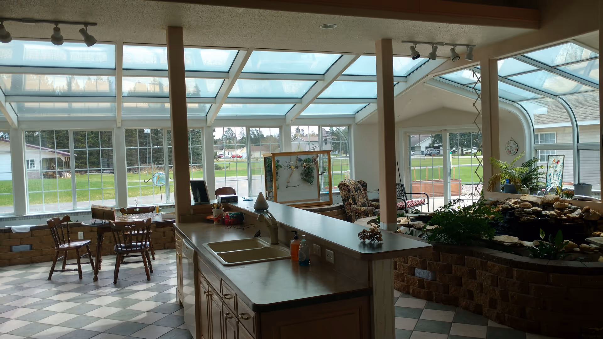 Interior view of a bright sunroom with large windows and a glass ceiling. The room features a kitchen counter with a sink, a small dining table with chairs, a seating area with armchairs, and a decorative indoor water feature surrounded by plants. Outside, green grass and neighboring houses are visible through the windows.