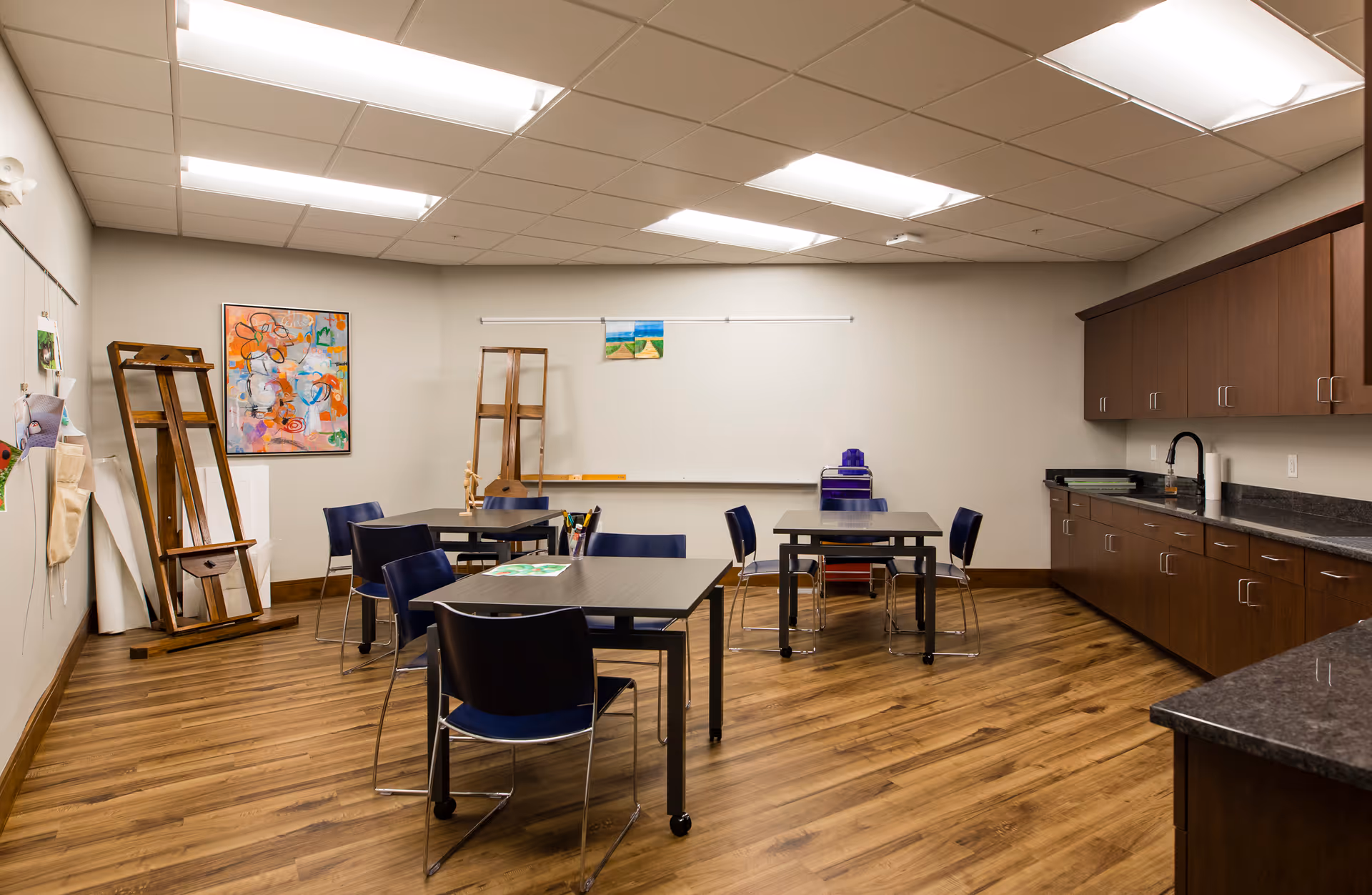 A well-lit room with wooden flooring featuring three tables each surrounded by blue chairs. The room has a whiteboard on the far wall, two wooden easels, and a colorful abstract painting. On the right side, there are dark wooden cabinets with a countertop and a sink. The ceiling has recessed lighting.