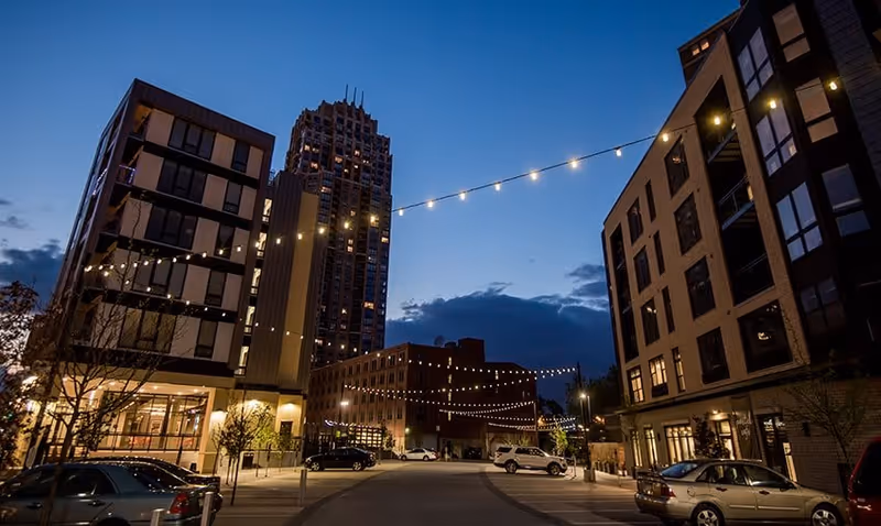 Evening view of a modern urban street scene with string lights hanging between buildings, several parked cars, and a tall skyscraper in the background under a darkening blue sky.