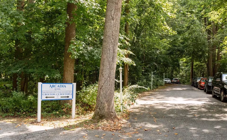 Tree-lined paved driveway with parked cars and an 'Arcadia Living' directional sign for Crofton at the entrance.