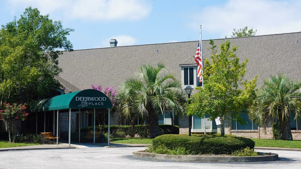 Exterior view of a single-story brick building with a green awning labeled 'Deerwood Place' over the entrance. The building is surrounded by palm trees, bushes, and other greenery. An American flag is flying on a flagpole near the entrance under a partly cloudy sky.