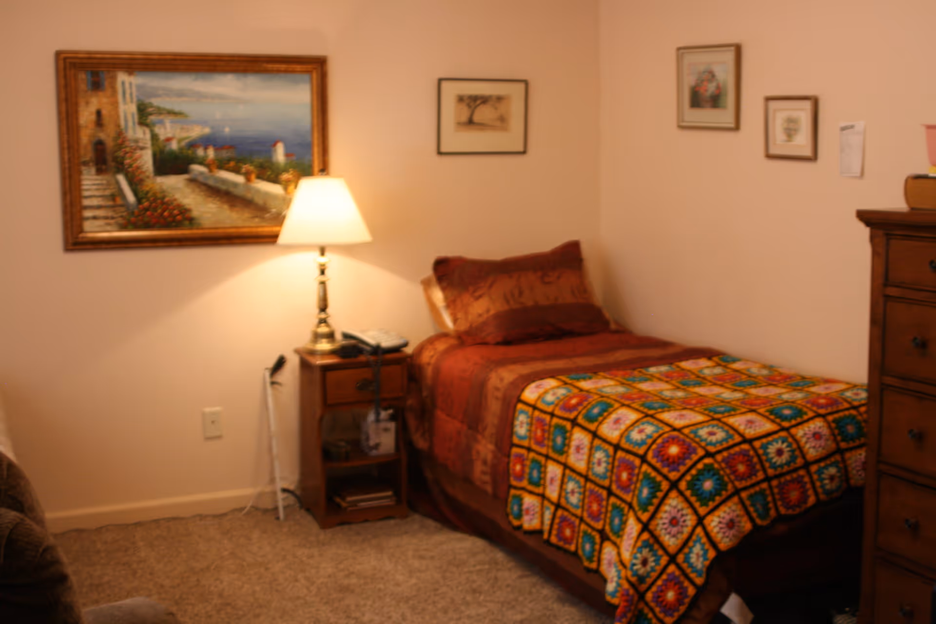 A cozy bedroom with a single bed covered in a brown and colorful crocheted blanket. Next to the bed is a wooden nightstand with a lamp, a telephone, and some items on the lower shelf. The walls are decorated with framed paintings and pictures. There is a wooden dresser on the right side of the image and a carpeted floor.