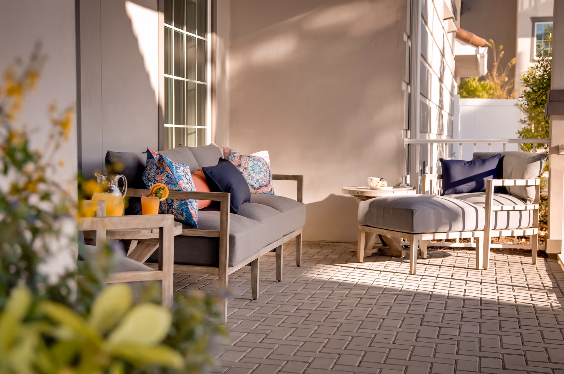 Outdoor patio area with cushioned wooden seating including a sofa and a chair with an ottoman, decorated with colorful pillows. A small table holds a pitcher of lemonade and a glass with a garnish. The patio has a tiled floor and is partially shaded with sunlight casting shadows on the wall.