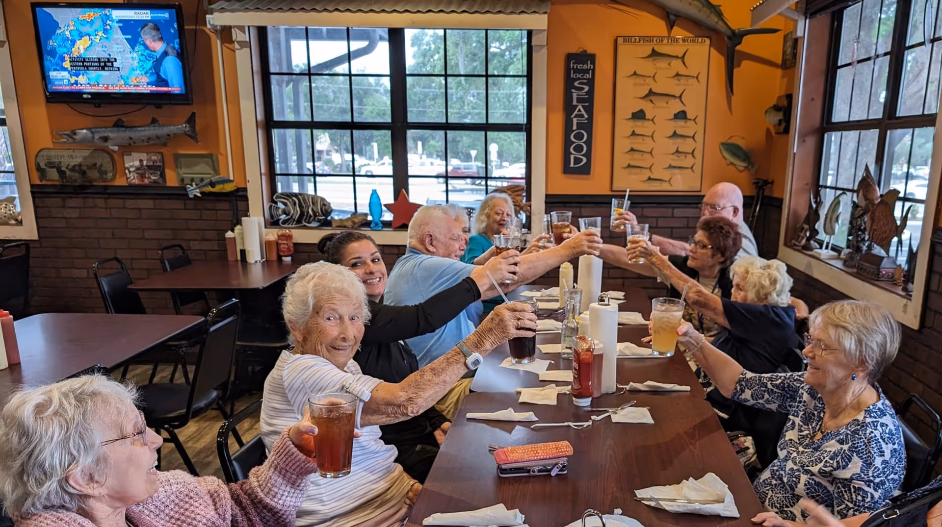 A group of elderly people and a caregiver sitting around a long dining table in a restaurant-style room, raising their glasses in a toast. The room has large windows, orange walls decorated with seafood-themed signs and fish models, and a television mounted on the wall.