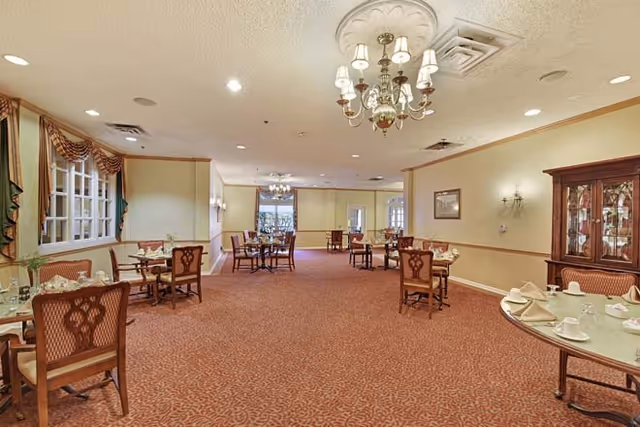 A spacious dining room with multiple tables and chairs arranged neatly. The room features warm lighting from chandeliers and wall sconces, a patterned carpet, and a china cabinet on the right side. Tables are set with white napkins and cups, and large windows with drapes are visible on the left wall.