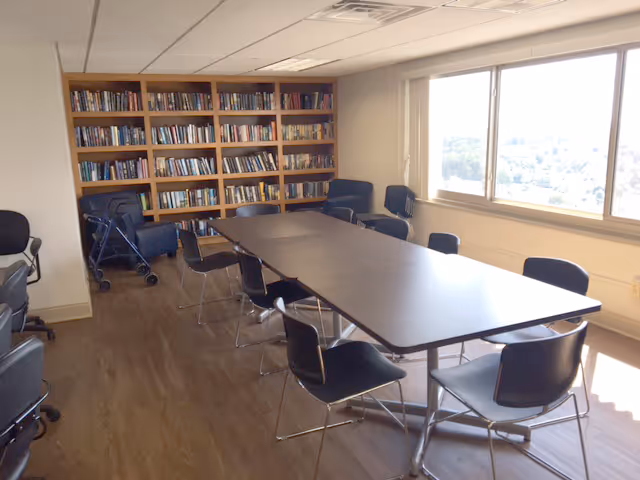 A meeting/reading room with a long table surrounded by chairs, bookshelves along the back wall, and a large window.