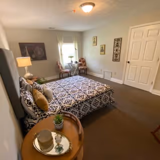 A cozy bedroom with a patterned bedspread on a bed, two chairs and a small table near a window with curtains, a lamp on a side table, and framed artwork on the walls.
