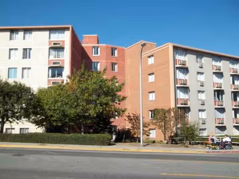 Exterior view of a multi-story retirement home with gray and tan facades and trees.