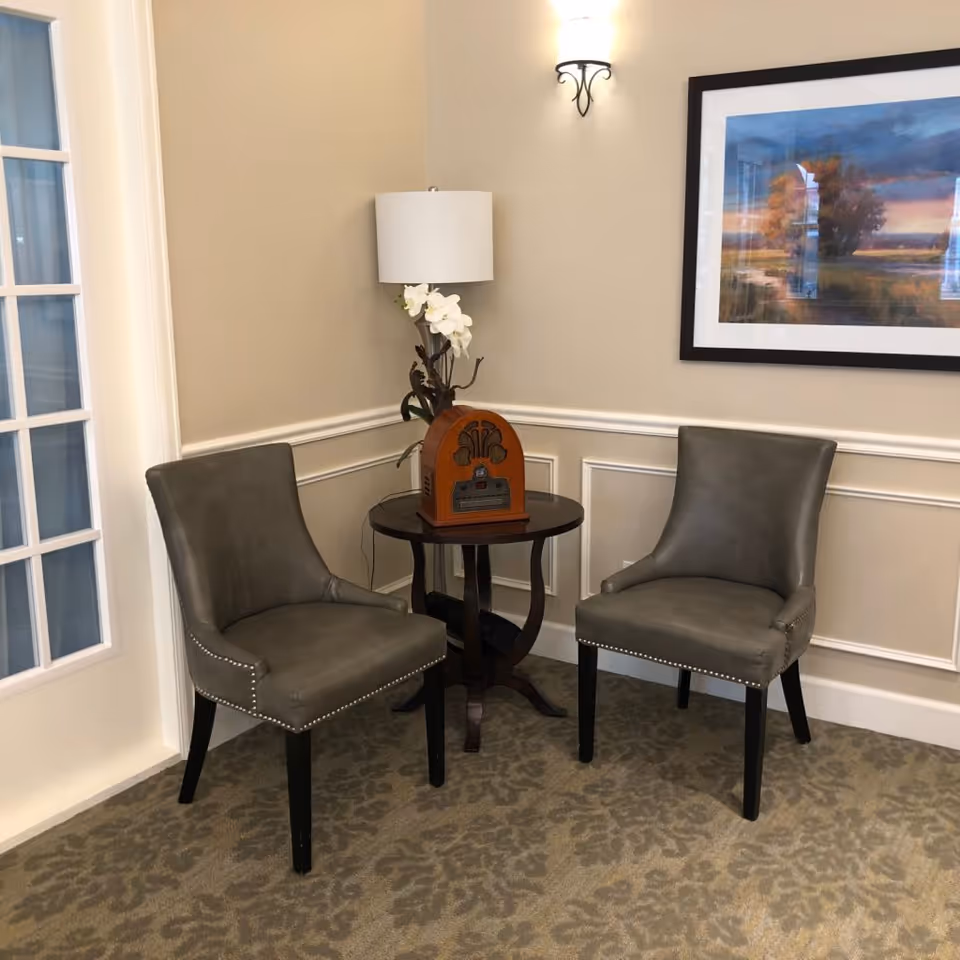 Two gray accent chairs flank a small round table holding a lamp, flowers and a decorative radio beneath framed artwork in a corner seating area.