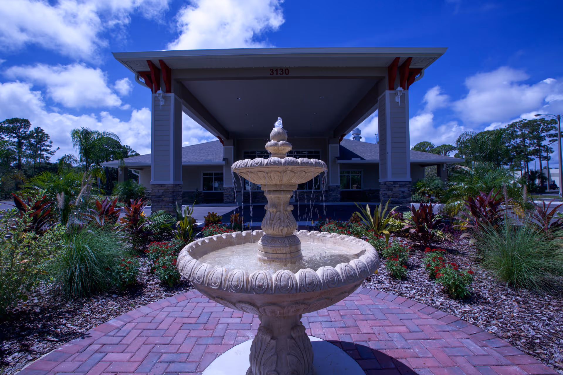 Outdoor view of the entrance to Majestic Gardens Living facility featuring a decorative two-tier stone water fountain in the foreground, surrounded by landscaped plants and flowers, with a covered driveway and building entrance in the background under a partly cloudy blue sky.