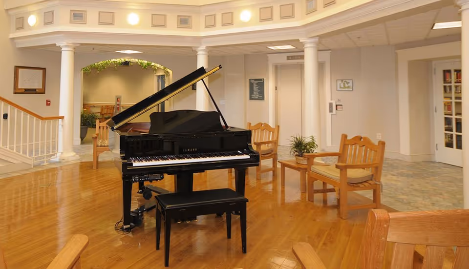 Interior common area with a black grand piano in the center, surrounded by wooden chairs with cushions. The room has wooden flooring, white columns, and soft lighting. There is a staircase on the left and an elevator in the background.