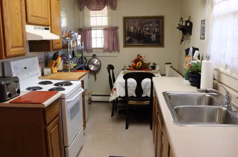 Small galley kitchen with a stove and wooden cabinets on the left, a double sink and counter on the right, and a small dining table with chairs at the far end.
