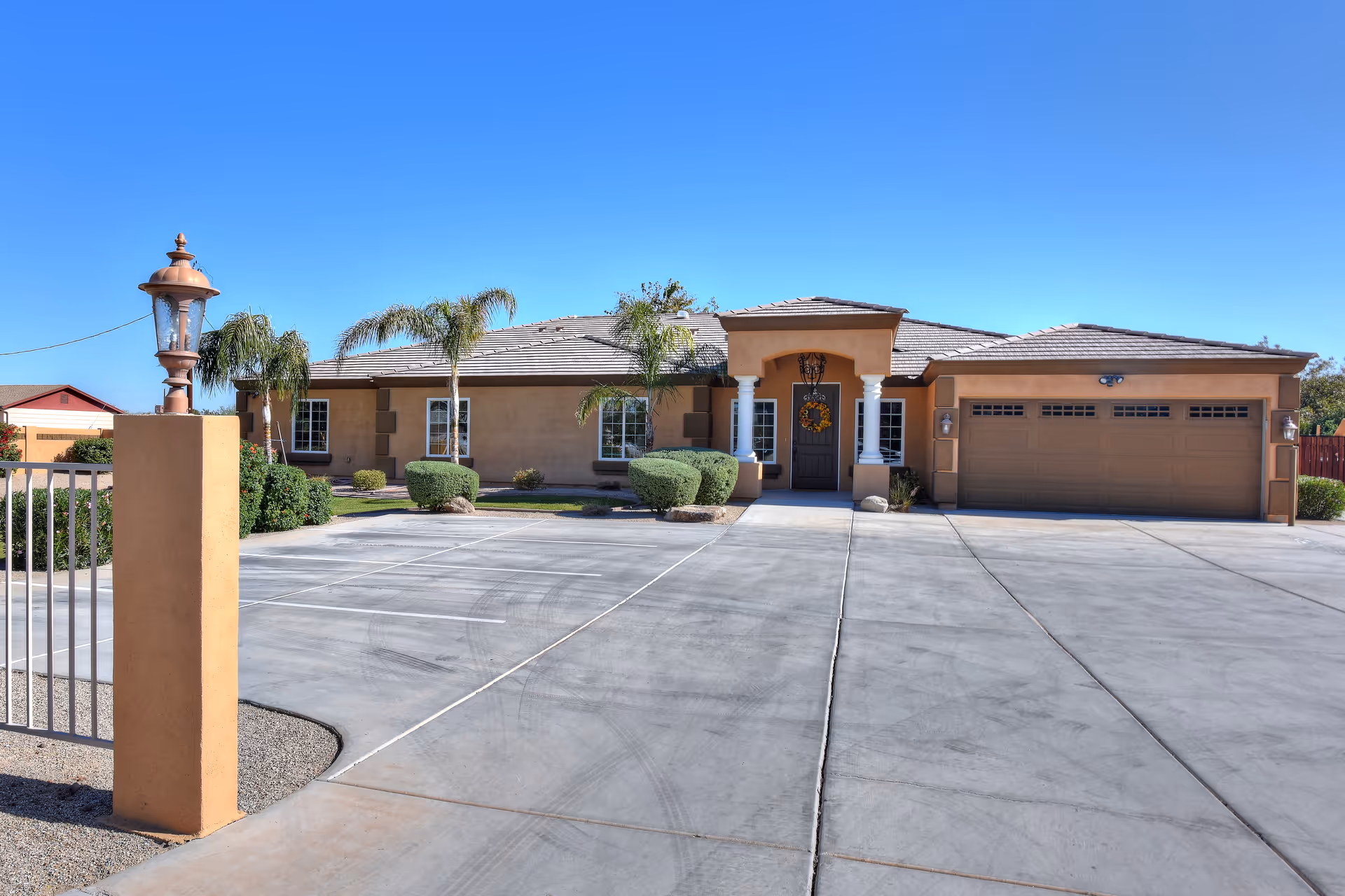 Front exterior view of a single-story house with a wide driveway, a garage, palm trees, and neatly trimmed bushes under a clear blue sky.
