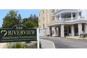Entrance view of Riverview Healthcare Community building with a sign in the foreground displaying the name and address, surrounded by greenery and a driveway leading to the building's covered entrance.
