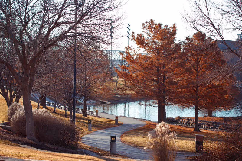 A winding paved pathway through a park-like area with leafless and autumn-colored trees on either side, leading towards a body of water with a bridge visible in the background under a clear sky.