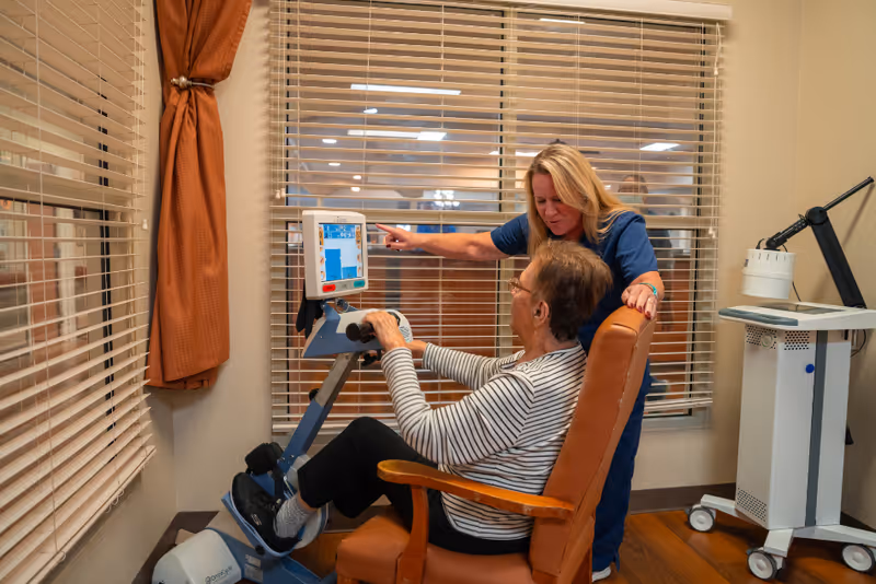 An elderly woman seated in a chair using a hand and foot exercise machine while a caregiver in blue scrubs stands beside her, pointing at the machine's screen. The room has beige walls, wooden flooring, and windows with blinds and rust-colored curtains.