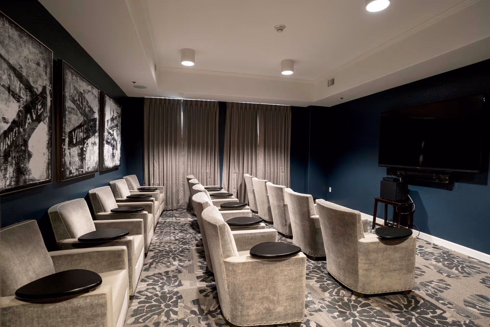 A small theater room with two rows of beige upholstered chairs, each equipped with a small black tray table. The walls are painted dark blue, and there are three black and white framed artworks on the left wall. A large flat-screen TV is mounted on the right wall, with a speaker and electronic equipment below it. The room has patterned carpet and curtains covering the windows at the back.