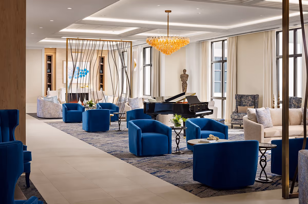 A spacious and elegant senior living facility common area featuring multiple blue armchairs arranged around small tables, a black grand piano, beige sofas, large windows with cream curtains, a modern chandelier, and decorative sculptures and artwork.