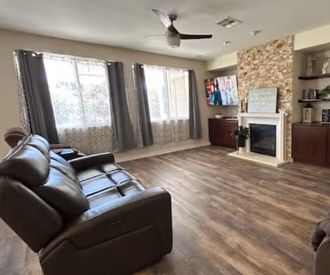 A spacious living room with wooden flooring, a dark brown leather reclining sofa, two large windows with patterned curtains, a ceiling fan, a stone fireplace with a white mantel, a mounted flat-screen TV, and built-in wooden shelves and cabinets on either side of the fireplace.