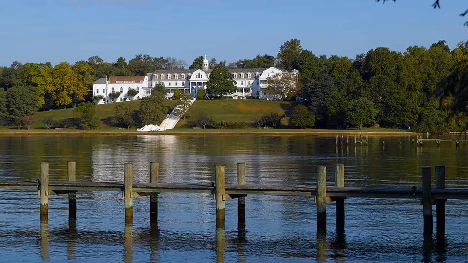 A large white building situated on a grassy hill surrounded by trees, with a wooden dock extending into a calm body of water in the foreground under a clear blue sky.