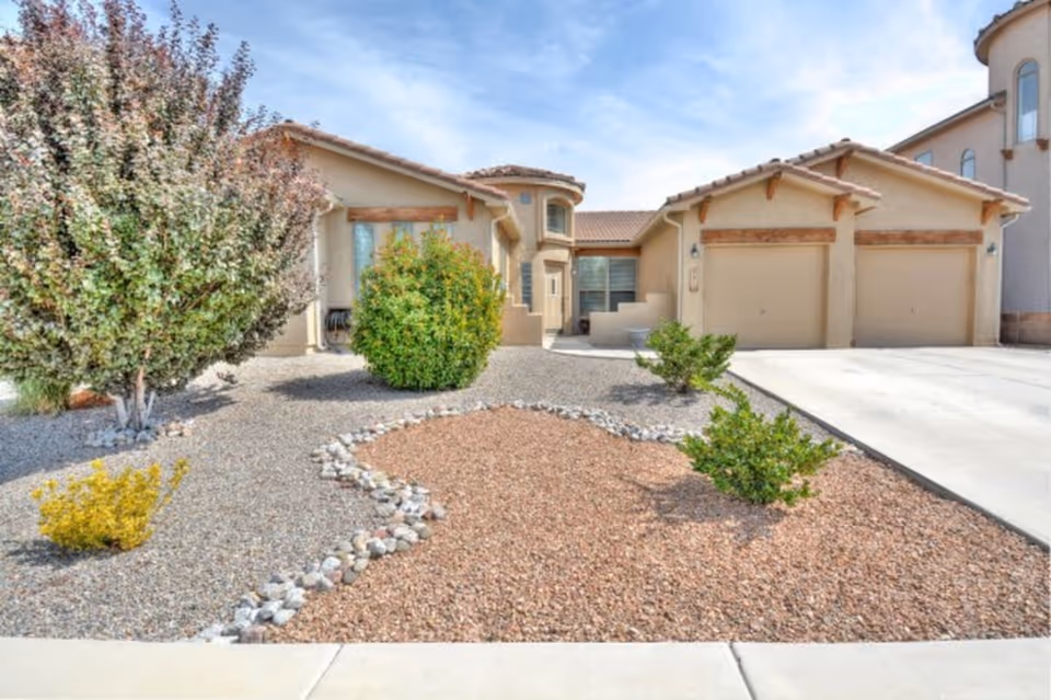 Front exterior of a single-story stucco house with a three-car garage and gravel xeriscaped yard with shrubs.