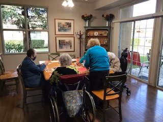 A group of elderly people sitting around a wooden table in a well-lit room with large windows. One person in a wheelchair is visible, and another person is standing, engaging with the group. The room has wooden floors, framed pictures on the walls, and a glass door leading to an outdoor patio area with chairs.