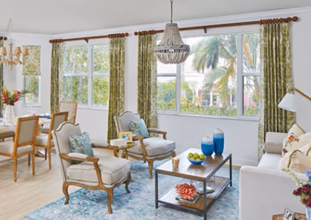 A bright and airy living room and dining area with large windows covered by green patterned curtains. The living room features two cushioned armchairs with wooden frames, a white sofa with floral pillows, a wooden coffee table with decorative items including blue vases and a bowl of green apples, and a blue patterned rug. The dining area has a wooden dining table with six chairs and a chandelier above it. The room has light wood flooring and white walls.