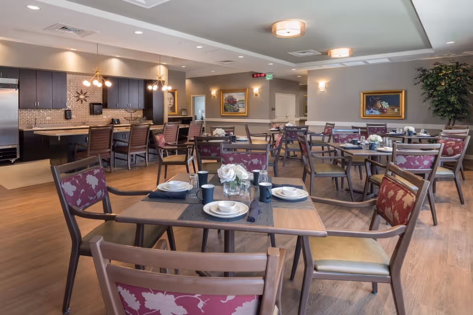 A spacious dining room in a senior living facility with multiple wooden tables and chairs, some with floral upholstery. Tables are set with plates, cups, and napkins. The room features a kitchen area with dark cabinets, a refrigerator, and modern pendant lighting. The walls are decorated with framed paintings and there is a large potted plant in the corner.