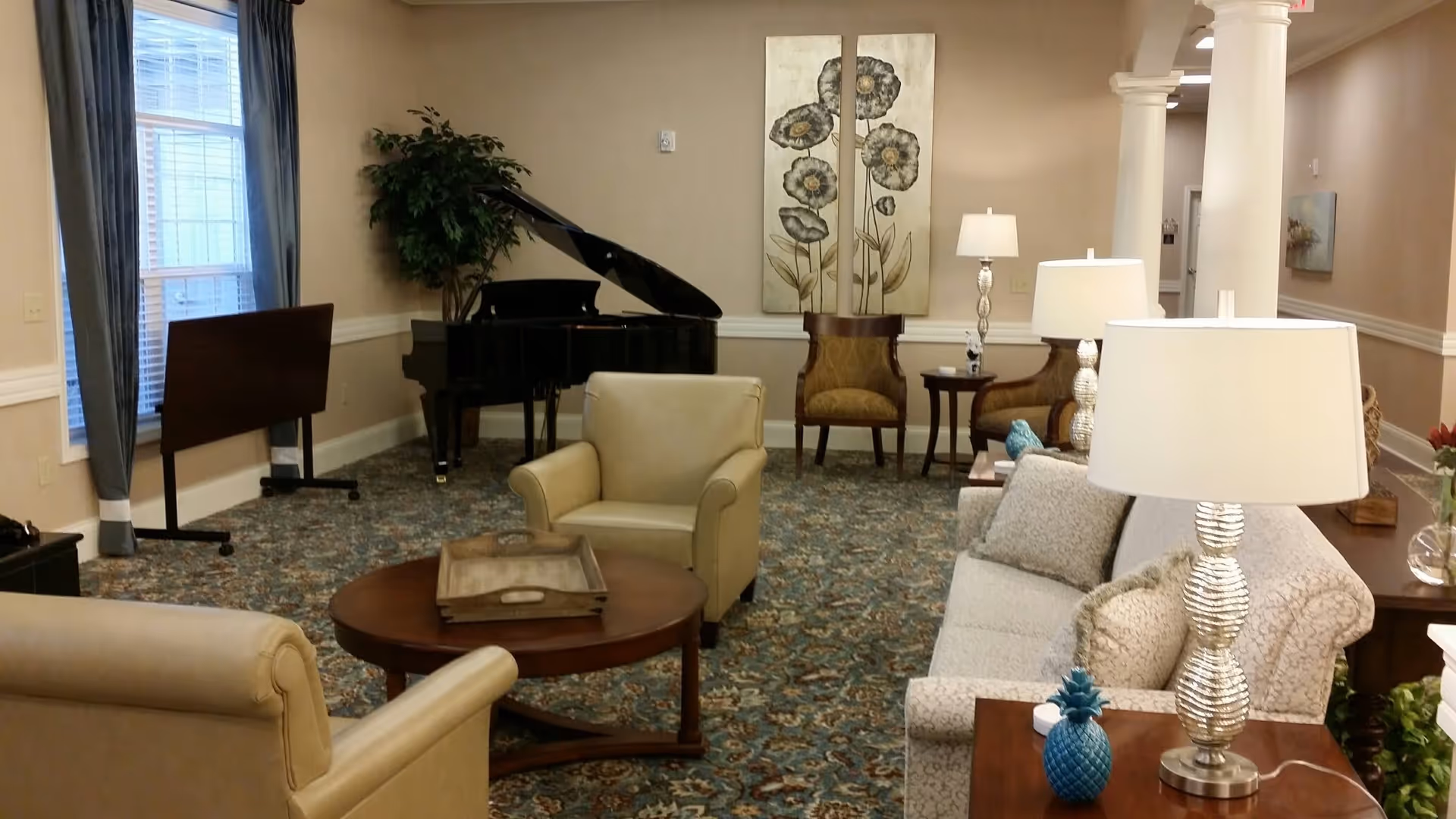 Communal lounge with sofas, armchairs, side tables and lamps, a coffee table, decorative artwork, and a grand piano near a window.