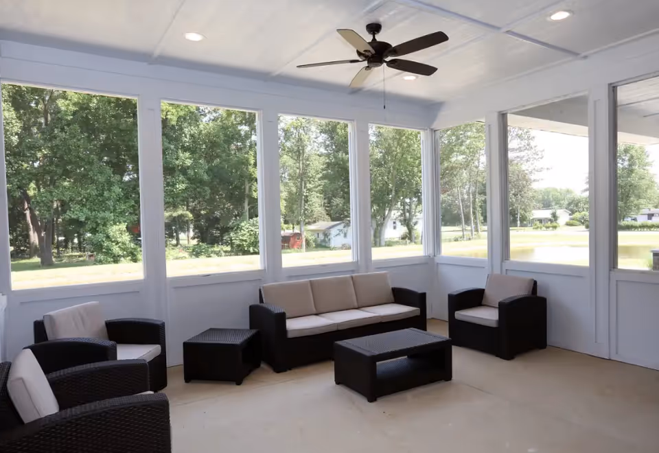 Bright screened-in sunroom with wicker sofa and chairs, a coffee table, ceiling fan, and large windows overlooking a lawn and trees.