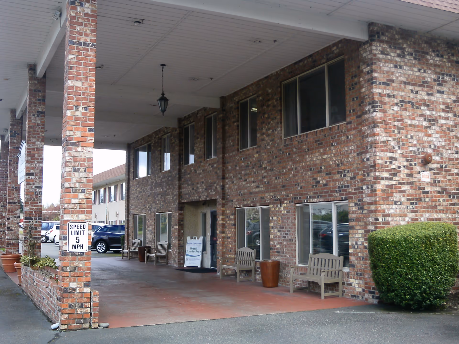 Covered entrance area of a brick building with several wooden benches and potted plants. There is a speed limit sign indicating 5 MPH and a sandwich board sign near the door advertising rent and smoke-free homes. Several windows are visible on the building, and cars are parked in the background.