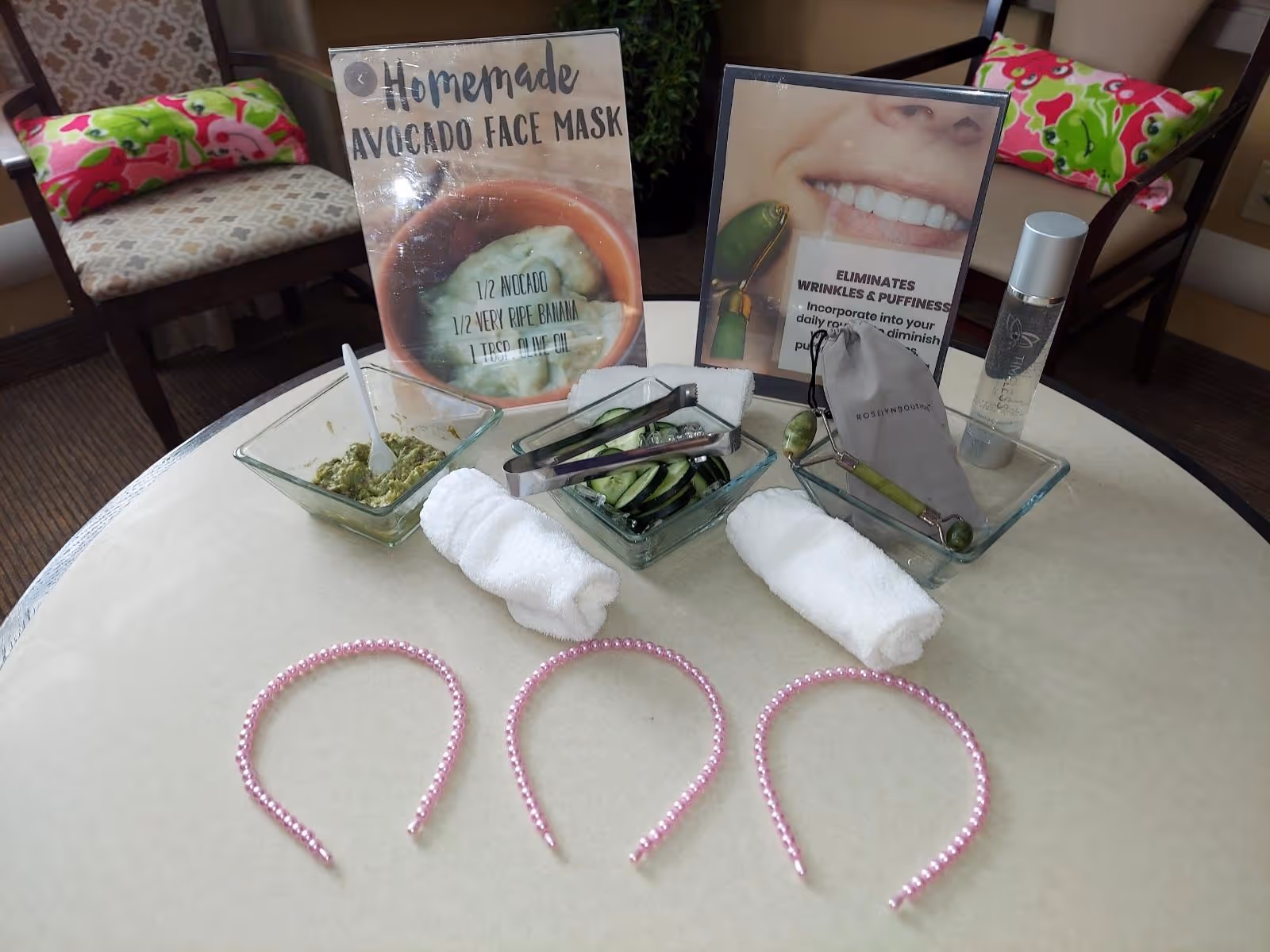 A table set up with homemade avocado face mask ingredients, cucumber slices, facial rollers, a bottle of skincare product, and three pink beaded headbands. Two signs on the table describe the avocado face mask recipe and the benefits of using a facial roller to eliminate wrinkles and puffiness. Two chairs with colorful cushions are visible in the background.