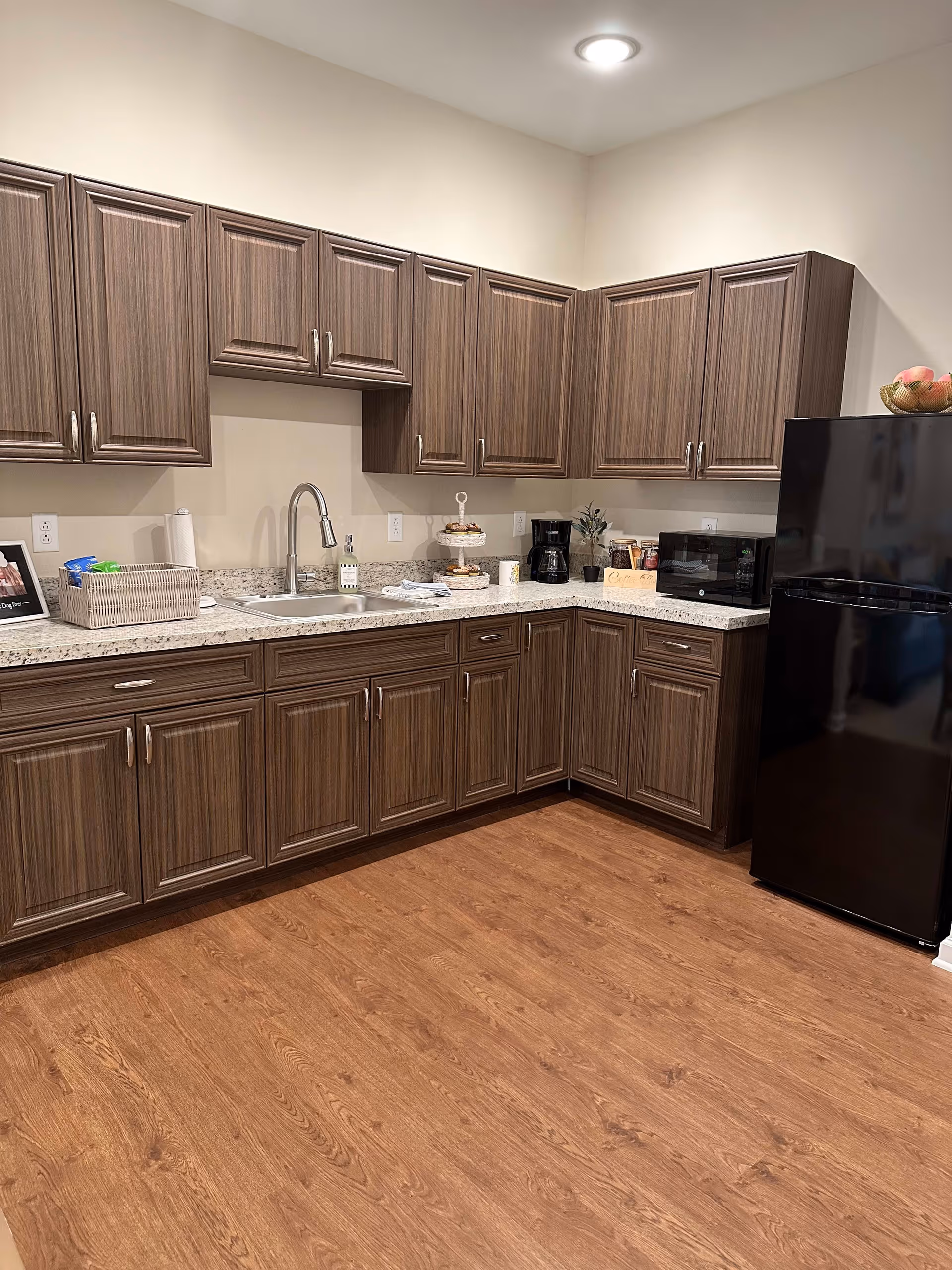 A kitchen area with wood grain cabinets, a granite countertop, a stainless steel sink with a modern faucet, a black refrigerator, a microwave, a coffee maker, and various kitchen items including a basket, paper towels, and a tiered tray with pastries. The floor is wood and the walls are painted light beige.