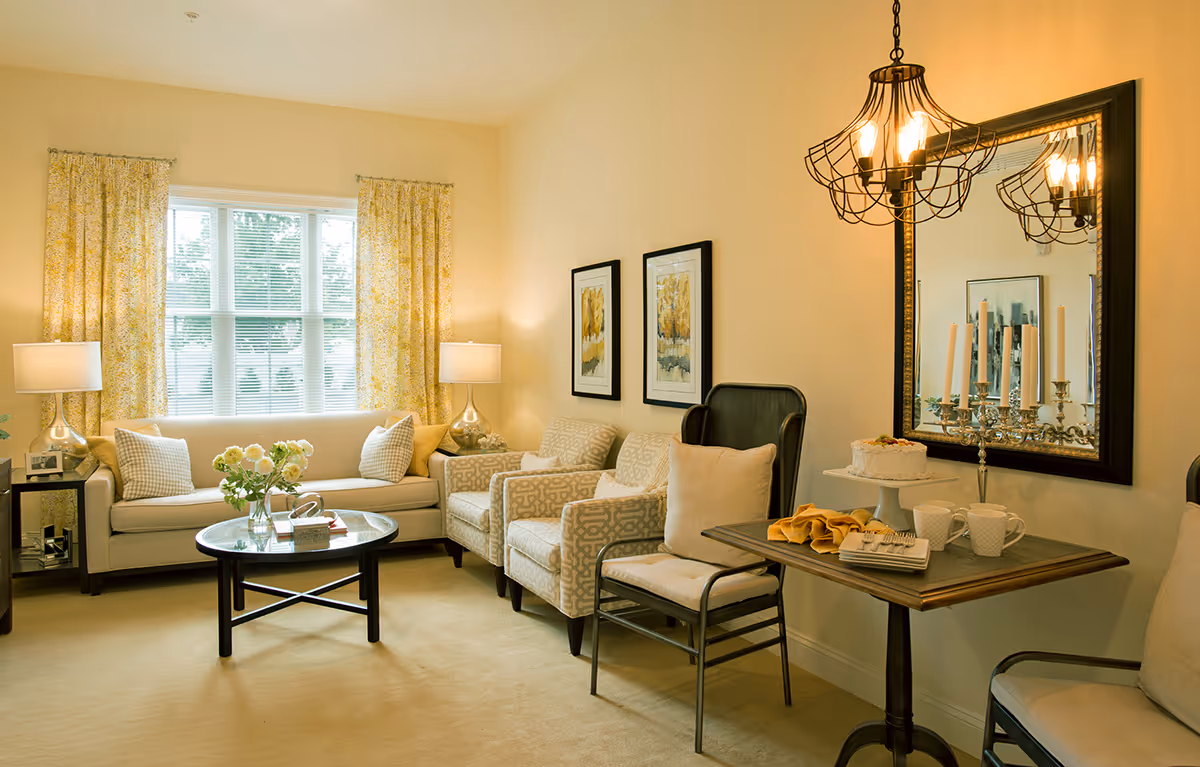 A cozy living room with a beige sofa adorned with pillows, two patterned armchairs, and a wooden chair with a cushion. A round glass coffee table with a flower vase sits in front of the sofa. The room features yellow patterned curtains on a large window, two framed artworks on the wall, and a large mirror above a small table. The table holds a cake on a stand, two mugs, napkins, and a silver candelabra. A decorative hanging light fixture is reflected in the mirror.