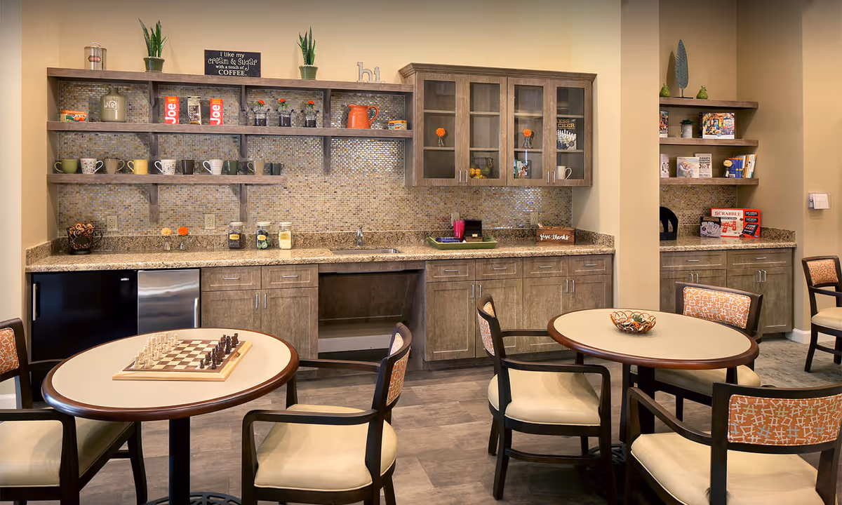 A cozy common area with round tables and chairs, one table has a chessboard set up. The background features a kitchenette with wooden cabinets, a mosaic tile backsplash, open shelves with mugs, jars, and decorative items. There are also board games on shelves to the right.