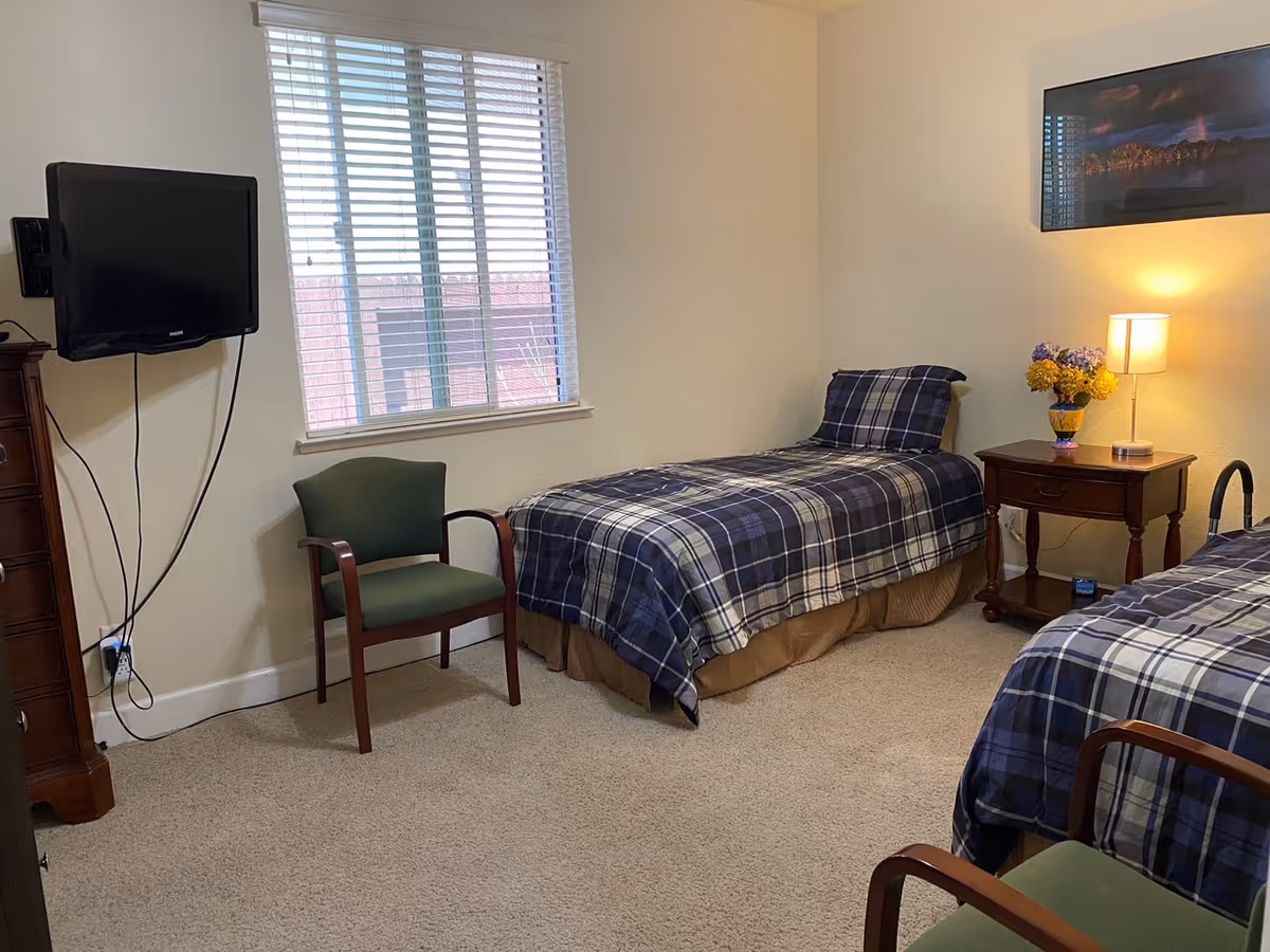 A tidy twin bedroom with two plaid single beds, a nightstand with lamp and flowers, a wall-mounted TV, and two chairs.
