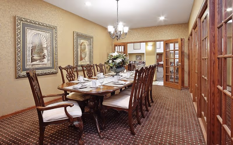 A formal dining room with a long wooden table set for eight people. The table is adorned with white plates, glasses, napkins, and a floral centerpiece. The room features patterned carpet, beige walls with framed artwork, and wooden French doors leading to another room.