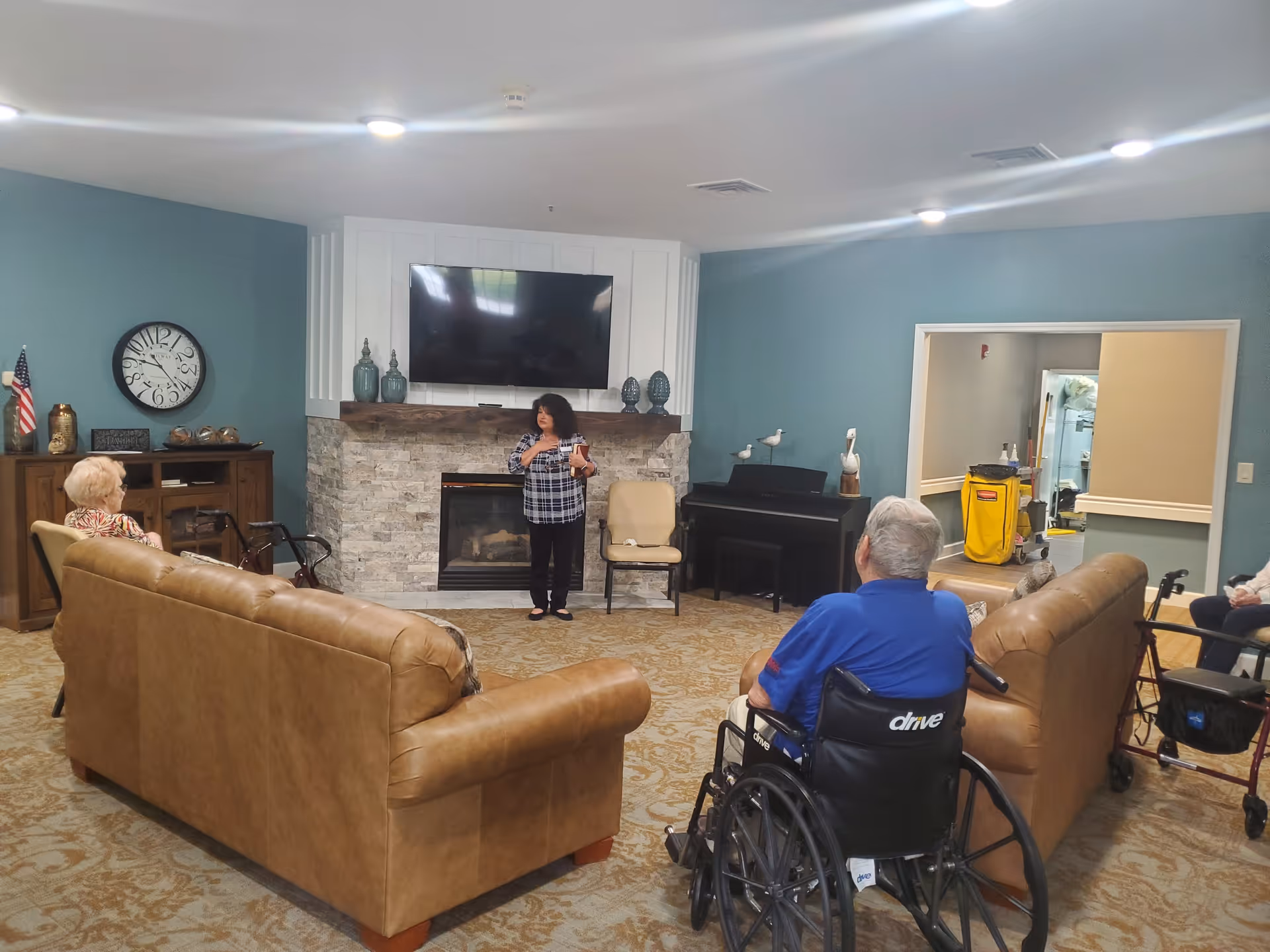 A common living room area in a senior living facility with two tan leather sofas facing a stone fireplace with a mounted flat-screen TV above it. A woman stands near the fireplace speaking to three elderly people seated around the room, one in a wheelchair and another with a walker. The walls are painted blue, and there is a wooden cabinet with a clock and American flag on the left side. A piano is visible against the right wall.