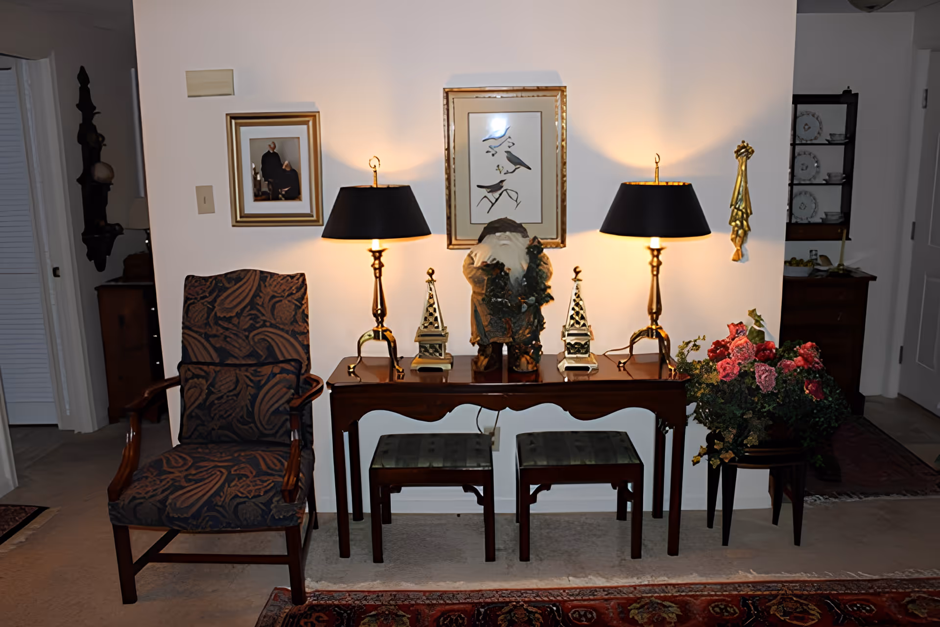 A cozy interior corner featuring a wooden armchair with patterned upholstery, a wooden console table with two black lamps, decorative figurines, and framed artwork on the wall. There are two small stools under the table and a floral arrangement on a stand to the right.