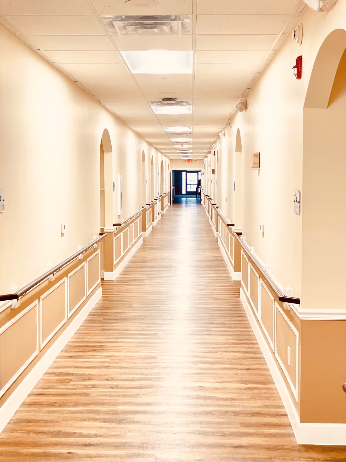 A long, well-lit hallway in a senior living facility with wooden flooring, beige walls, handrails on both sides, and several doorways along the corridor leading to rooms. The ceiling has recessed lighting and ventilation panels, and there is a door with windows at the far end of the hallway.