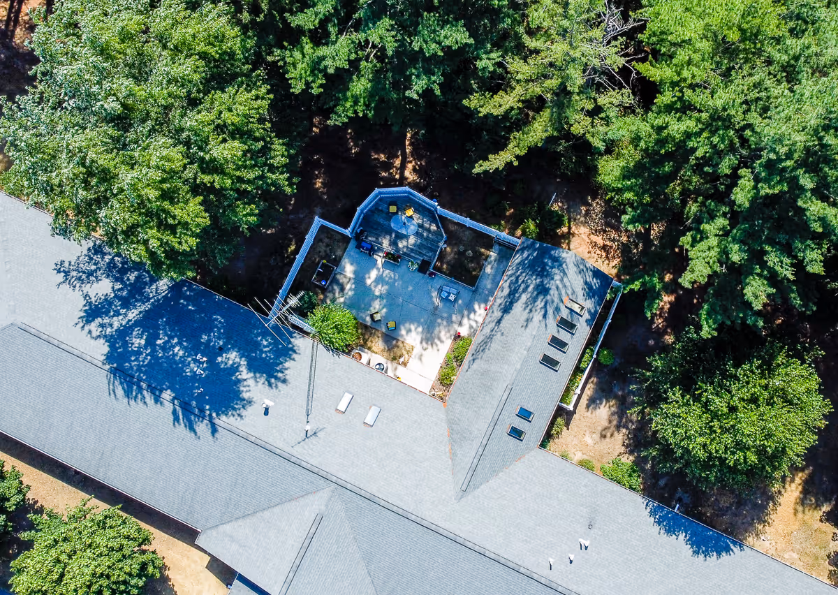 Aerial view of a senior living facility surrounded by dense green trees. The image shows the roof of the building with a fenced outdoor patio area containing seating and tables.