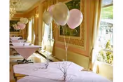 A bright dining room with tables covered in light pink tablecloths, each decorated with a small centerpiece and a cluster of pink and white balloons. Large windows with yellow curtains allow natural light to fill the room.
