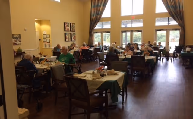 A dining room in a senior living facility with several elderly residents seated at tables covered with tablecloths, eating and socializing. Large windows with curtains allow natural light to fill the room, and the floor is wooden.
