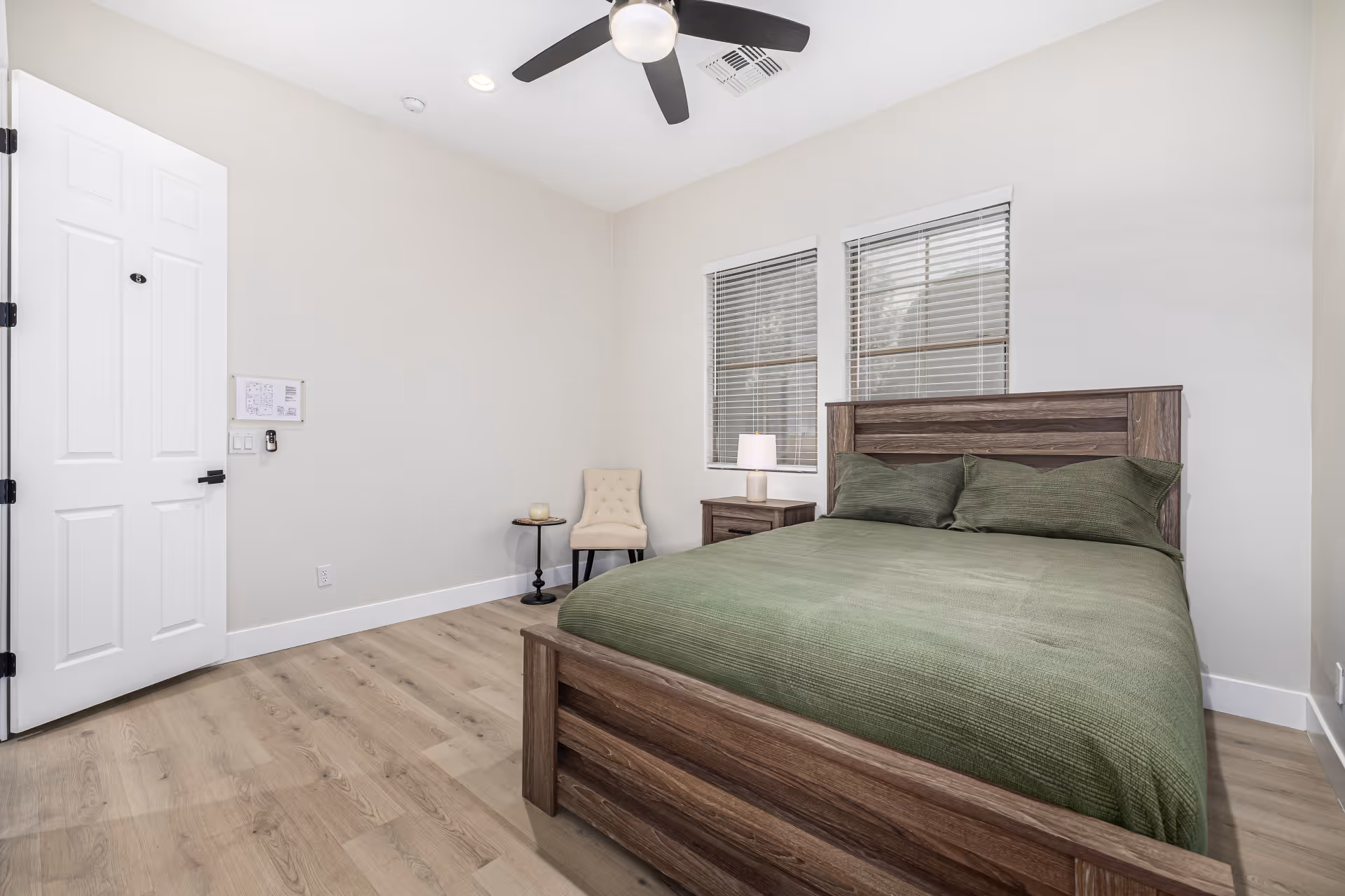 A bedroom with a wooden bed frame and green bedding, two windows with blinds, a nightstand with a lamp, a beige upholstered chair, and a small round side table. The room has light-colored walls, wood flooring, a ceiling fan with a light, and a white door.