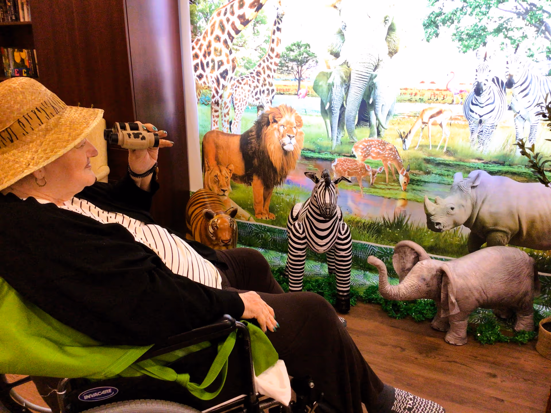 An elderly woman wearing a straw hat and black cardigan sits in a wheelchair looking through binoculars at a colorful wall mural and life-sized animal figurines including a lion, zebra, elephant, tiger, and rhinoceros in a room.