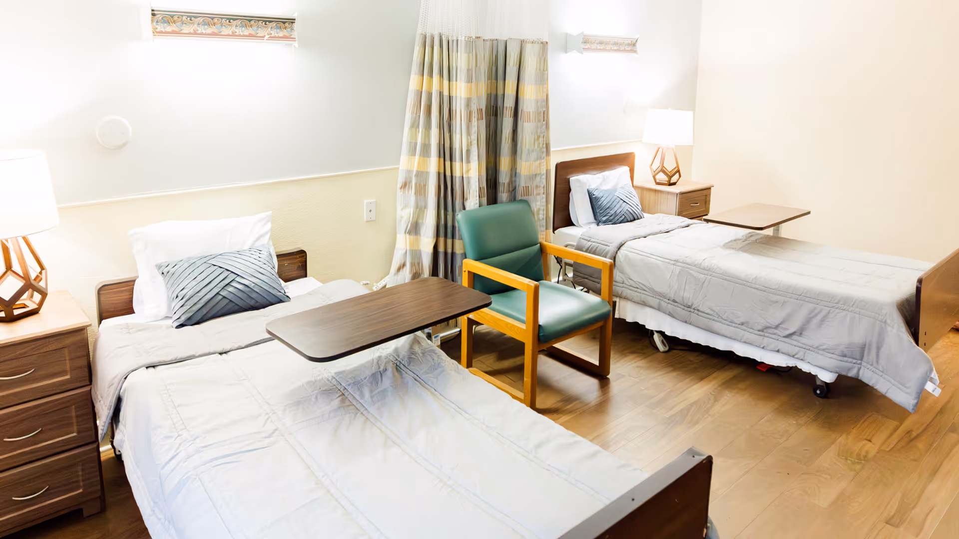 A bright room with two single beds, each with a gray comforter and a decorative pillow. Between the beds is a green armchair with wooden armrests. Each bed has a wooden bedside table with a lamp. There are curtains on the wall behind the beds and the floor is wooden.