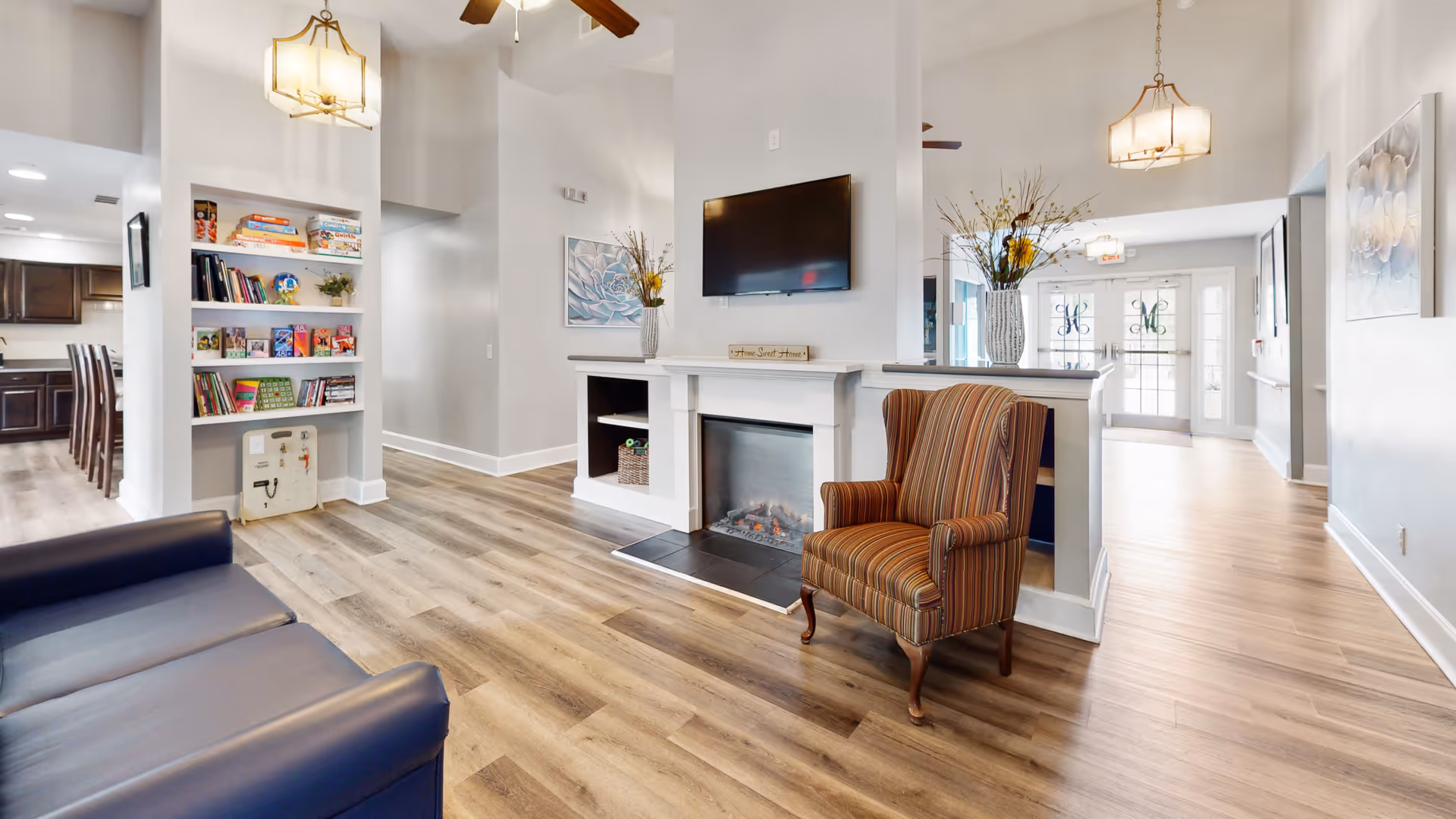 A bright and spacious living room area in a senior living facility with wood flooring, a striped armchair, a blue couch, a fireplace with a mounted TV above it, built-in shelves with books and games, and decorative vases with flowers. The room has high ceilings with ceiling fans and pendant lights, and large windows near the entrance letting in natural light.