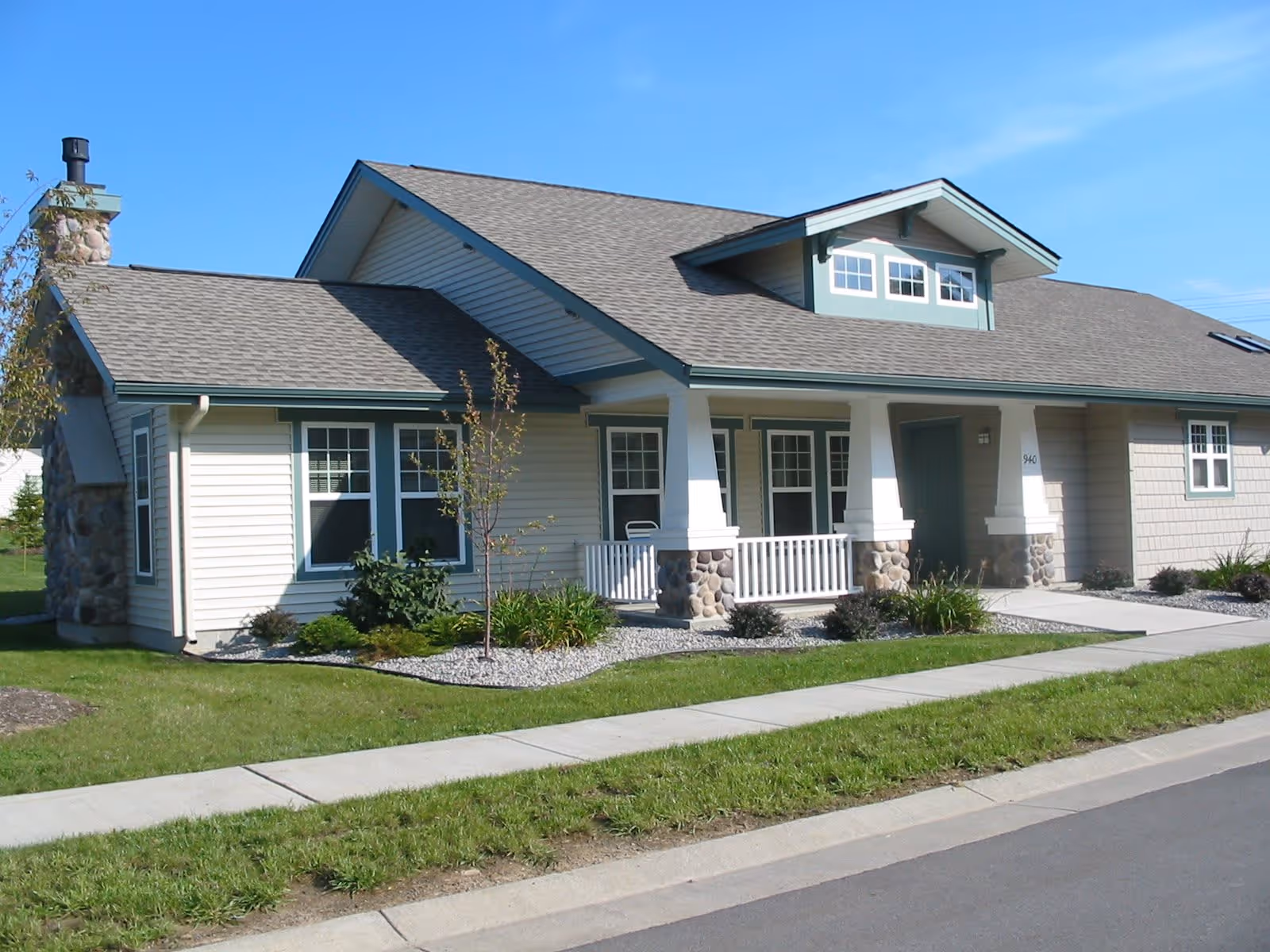 Exterior view of a single-story residential building with beige siding, green trim, a stone chimney, and a covered front porch with white railings. The building is surrounded by a well-maintained lawn, small shrubs, and a sidewalk in front.