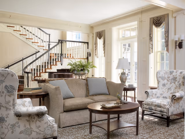 A bright and elegant living room with a beige sofa adorned with blue cushions, two floral-patterned armchairs, a wooden coffee table with decorative items, a side table with a lamp, and a staircase with black railings in the background. Large windows with drapes allow natural light to fill the space.